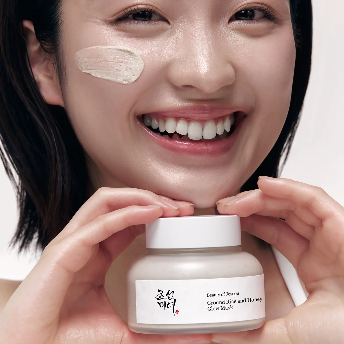 Beauty of Joseon Ground Rice and Honey Glow Mask model holding jar with mask on cheek